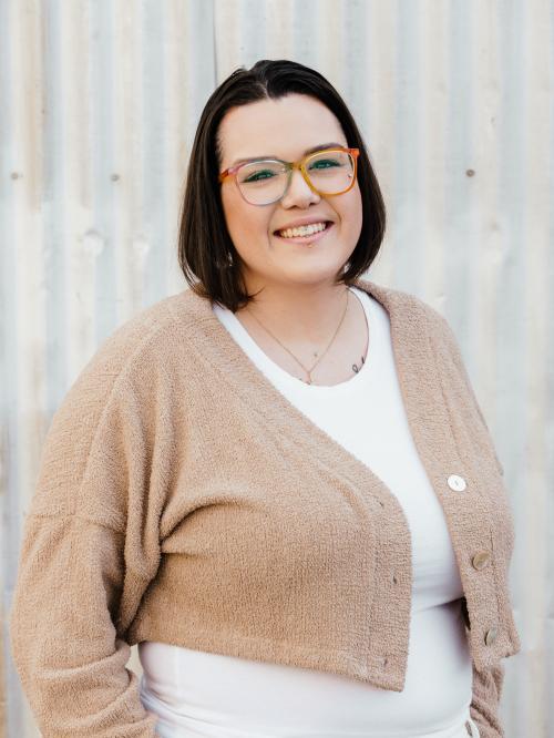 Pia Wright smiling, wearing colorful glasses and a tan cardigan.