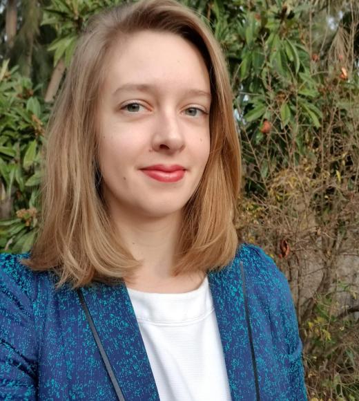 [alt text] headshot of Prof. Delmolino wearing a blue blazer with green foliage in the background