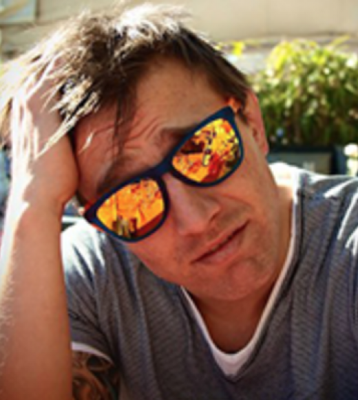 A man with his hand on his head. He is wearing sunglasses with orange lenses