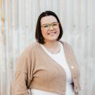 Pia Wright smiling, wearing colorful glasses and a tan cardigan.