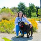 Lauren Watson, wearing a white lab coat and blue scrubs, smiles for the camera with a large black dog with its tongue hanging out.