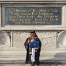 A woman in graduation regalia stands before a dedication for Yale university