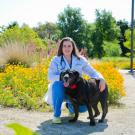 Lauren Watson smiles for the camera, wearing a white lab coat and blue scrubs. She has her arm over a large black dog with its tongue hanging out.