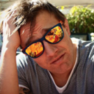 A man with his hand on his head. He is wearing sunglasses with orange lenses