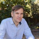Man in pale blue shirt, posing in front of greenery